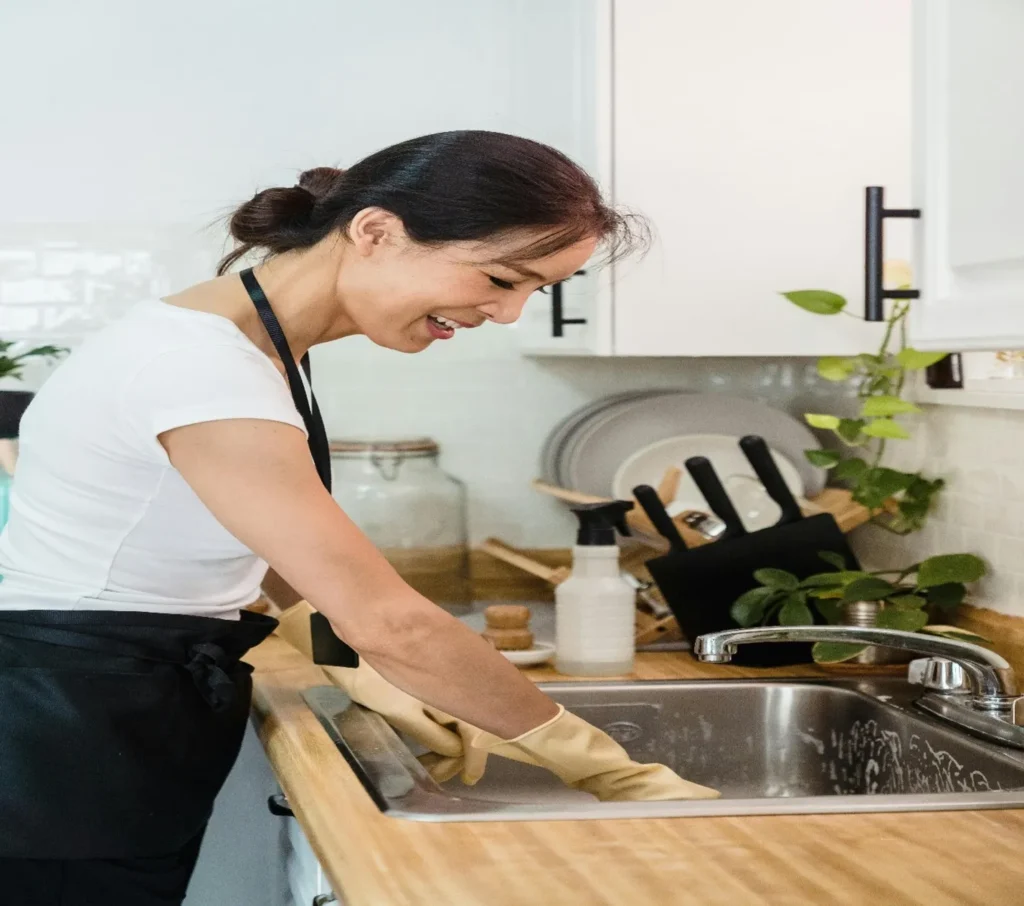 A woman in a white shirt is washing a kitchen sink with a sponge, surrounded by cleaning supplies.