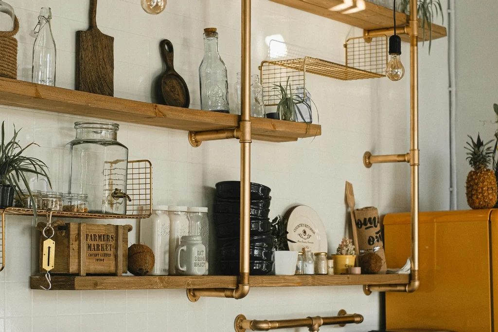 A cozy kitchen featuring a wooden shelf adorned with kitchen items and visible pipes along the wall.