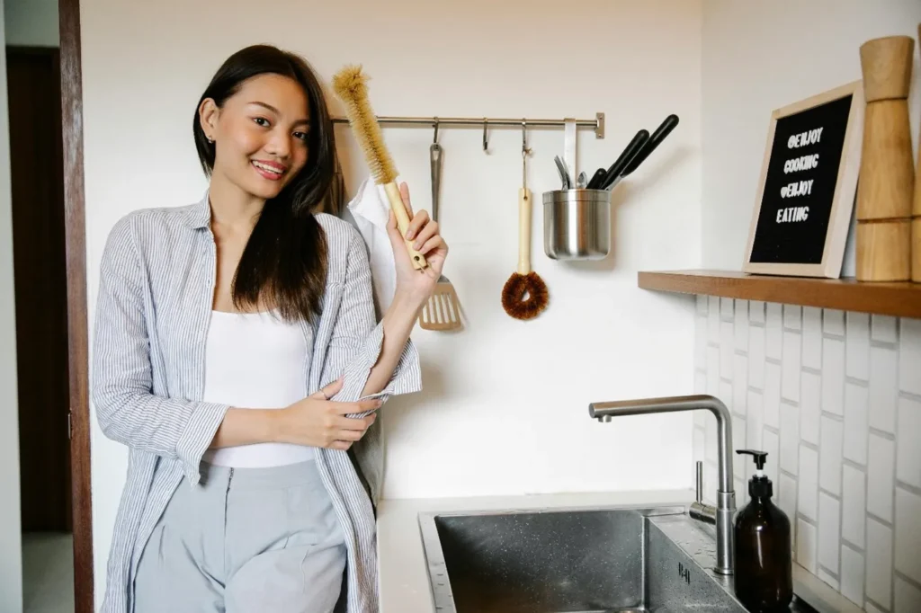 A woman standing in a kitchen holding a brush for cleaning kitchen sink pipes.