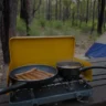 A cozy camping scene with a stove cooking sausages and soup on a picnic table in a forest, with a tent in view.