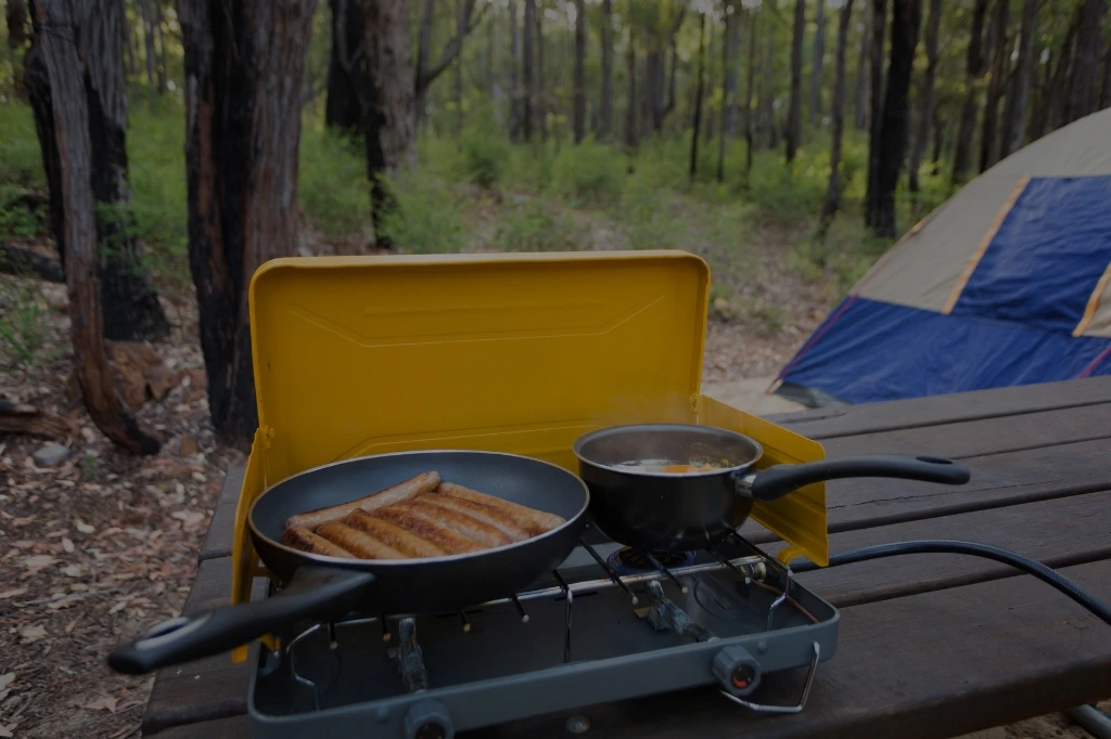 A cozy camping scene with a stove cooking sausages and soup on a picnic table in a forest, with a tent in view.