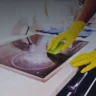 Gloved person scrubbing a glass stovetop with a sponge, with a blurred kitchen background of countertops and cabinets.