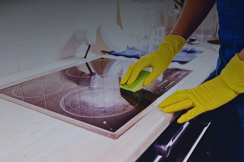 Gloved person scrubbing a glass stovetop with a sponge, with a blurred kitchen background of countertops and cabinets.