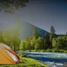 Orange tent by a blue river in a forested area, depicting potential summer cooking camp environment.