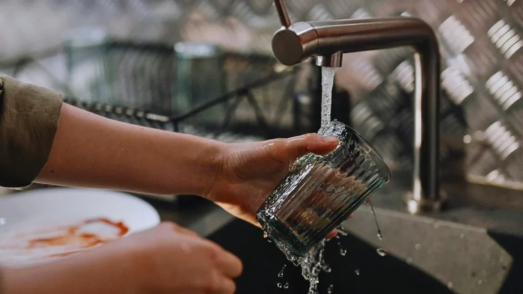 A person washing a glass under running water at a campsite, demonstrating how to clean kitchen equipment while camping.