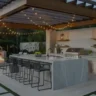 An elegant, modern outdoor kitchen layout ideas under a wooden pergola, featuring a large central island with built-in stainless steel appliances, a marble countertop, and stylish bar stools.