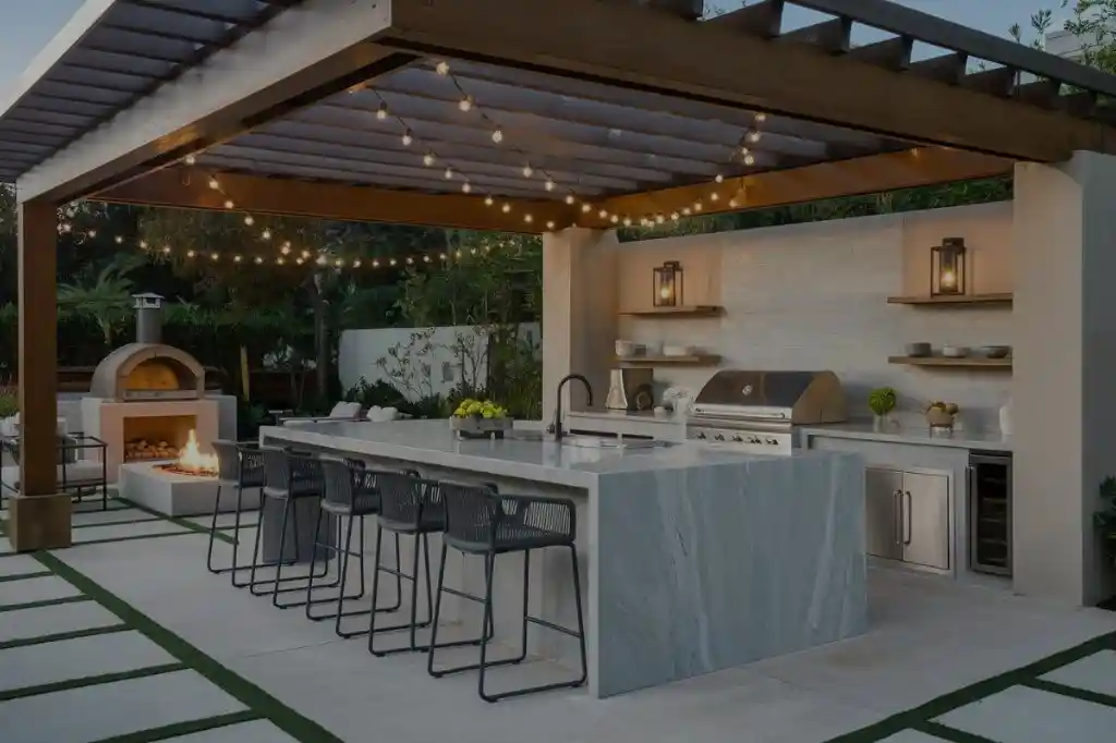 An elegant, modern outdoor kitchen layout ideas under a wooden pergola, featuring a large central island with built-in stainless steel appliances, a marble countertop, and stylish bar stools.