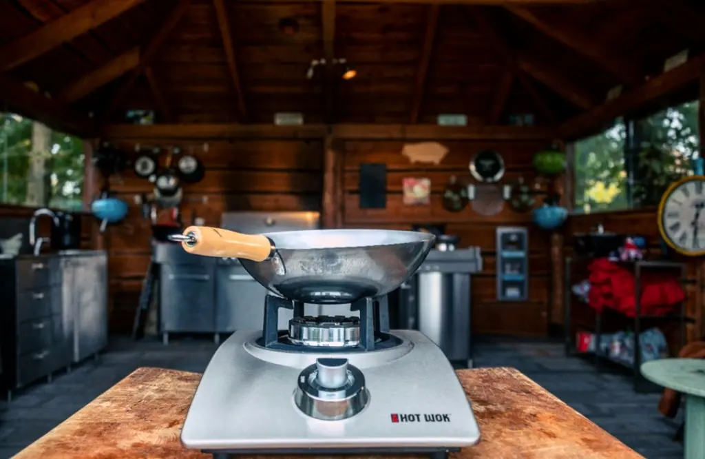 A portable gas stove with a wok on top, set in a wooden outdoor kitchen with various cooking utensils and appliances in the background.