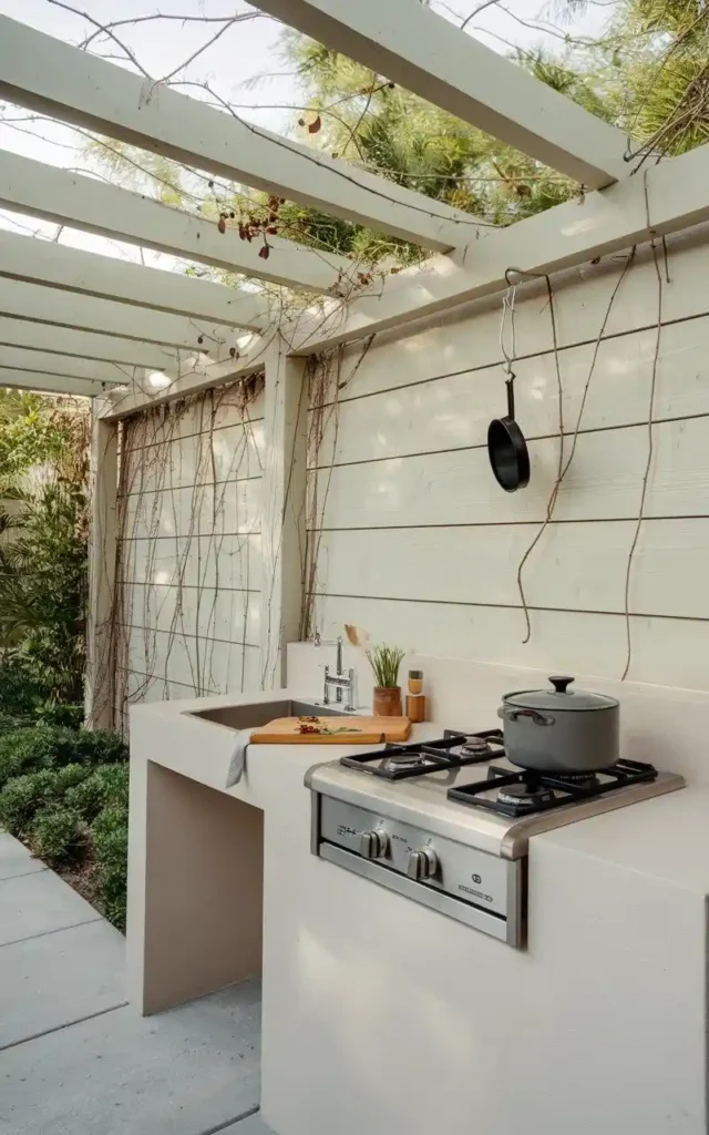 A sleek outdoor kitchen with a built-in counter, stove, and sink, surrounded by natural greenery under a pergola.