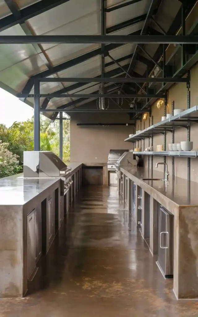 Galley-style outdoor kitchen with metal countertops, built-in grills, storage shelves, and industrial design under a covered steel structure.