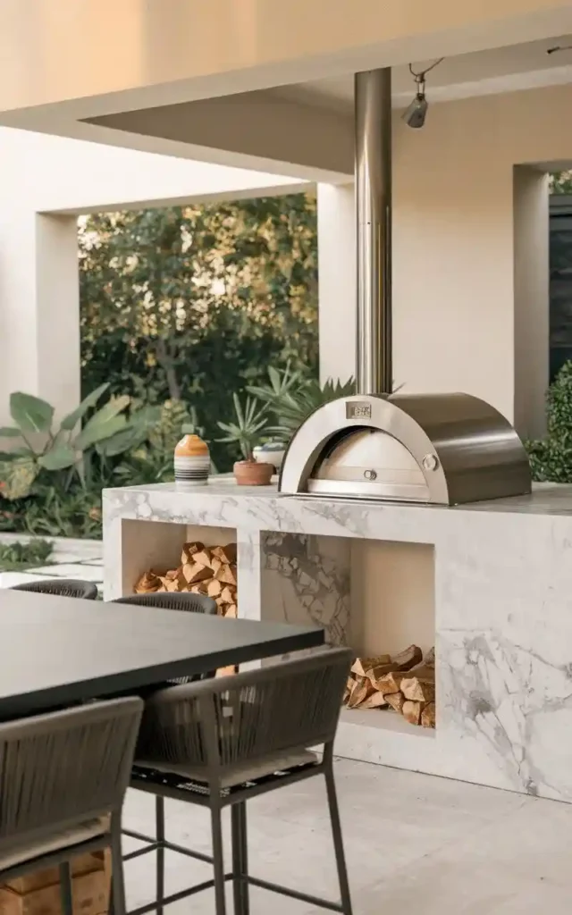 Outdoor kitchen with marble countertops, a stainless steel pizza oven, firewood storage, and a modern dining table, perfect for pizza parties.