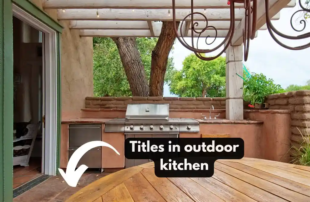 Outdoor kitchen with stainless steel grill, tiled countertops, and wooden dining table under a pergola.