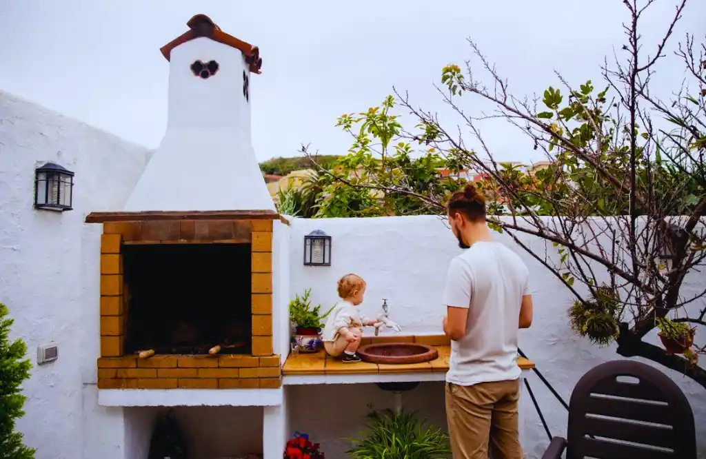 A man and a child at an outdoor kitchen sink station next to a traditional brick grill, with white walls and a garden with trees surrounding the area.